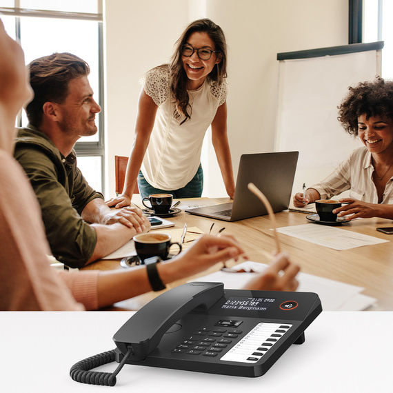 Modern desk phone with display and function keys, in the background a group in a meeting.