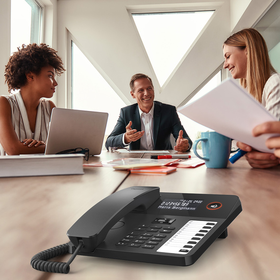 Modern desk phone with display and function keys, in the background a group in a meeting.